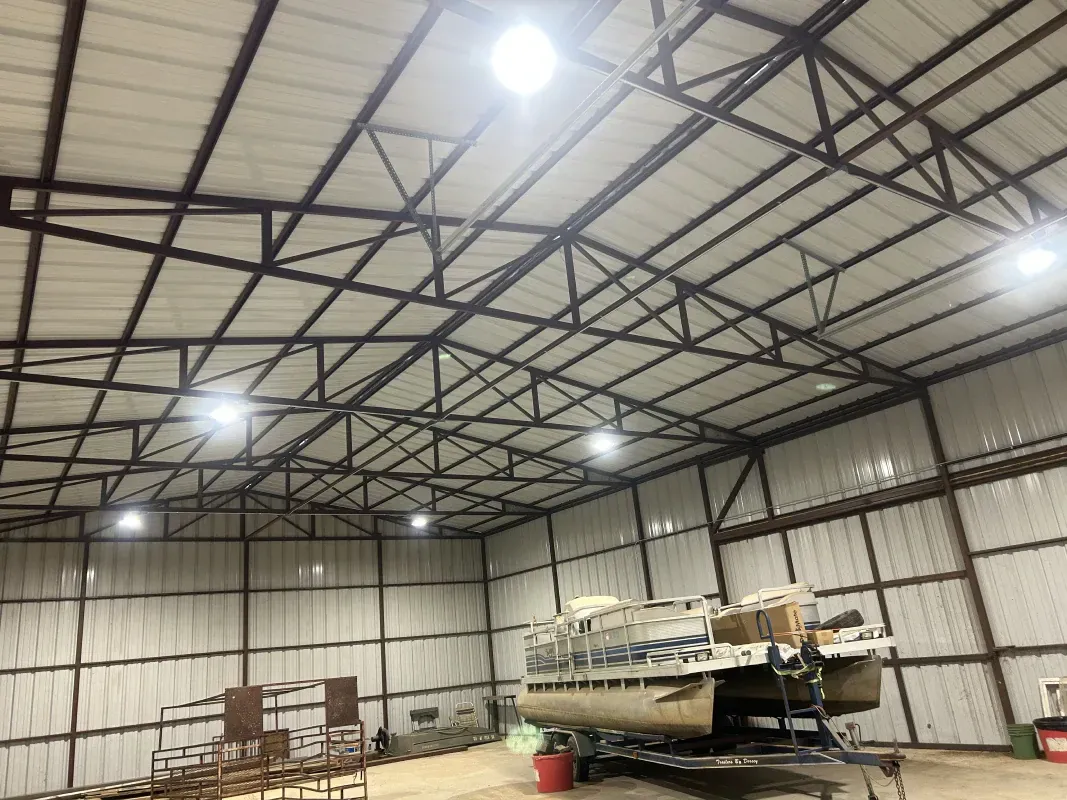 Inside a large metal building, a pontoon boat sits on a trailer. Overhead, bright lights illuminate the space.