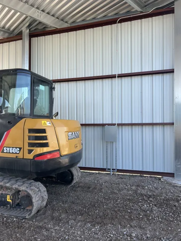 Yellow excavator next to a corrugated metal building with electrical box; gravel ground.