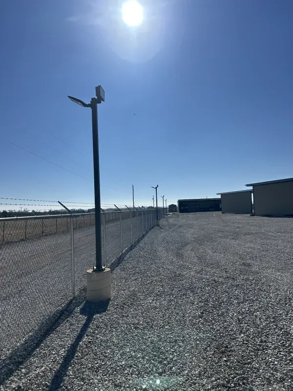 Chain link fence, gravel lot, blue sky with bright sun. Dark pole with light and camera. Buildings in background.