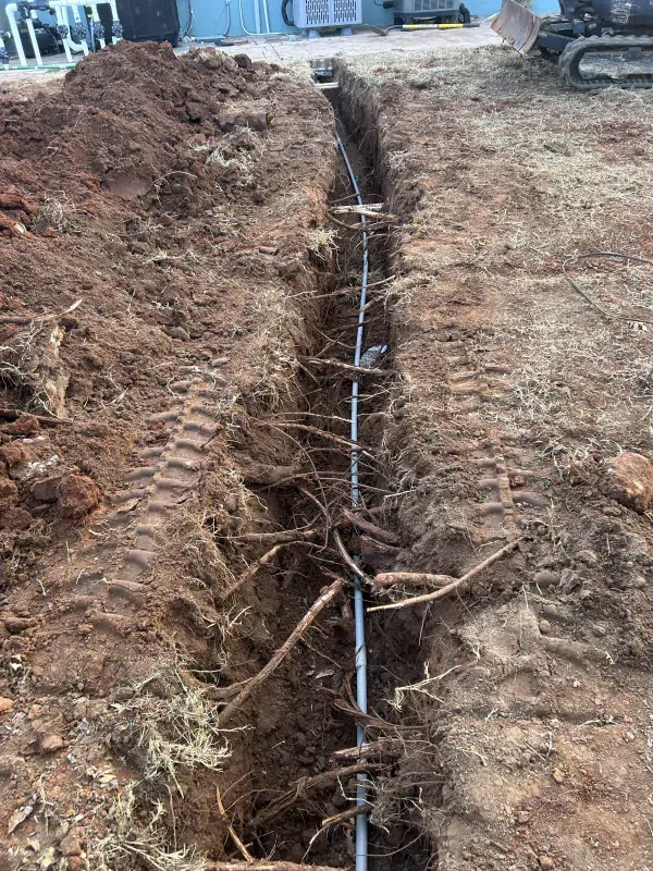 Trench dug in dirt with a silver pipe, roots, and tire tracks present.