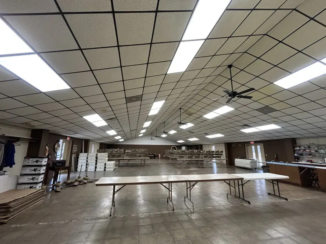 Large, empty room with long tables, fluorescent lighting, and a ceiling fan.