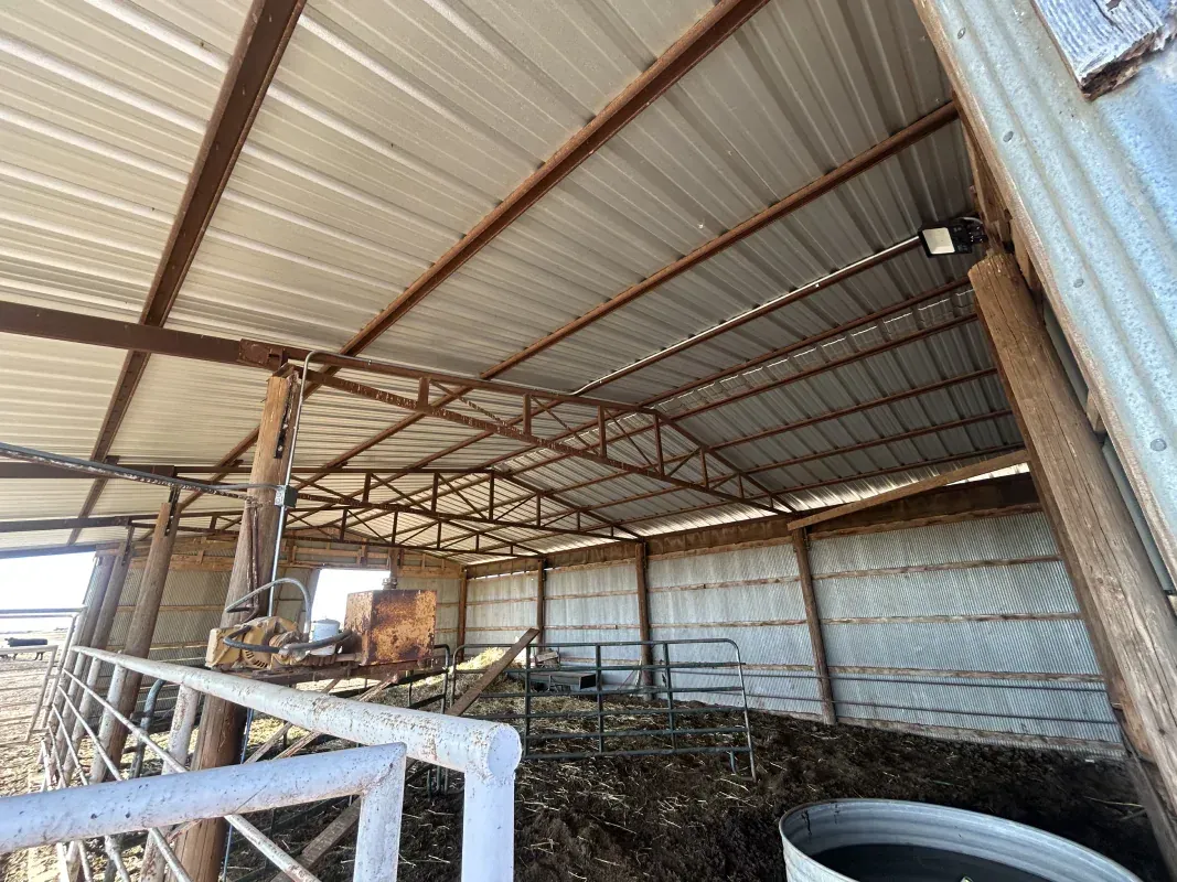 Open-air barn with metal roof and rusted supports; livestock area with hay and water trough.
