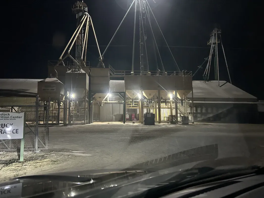 Grain elevator at night, illuminated by bright lights. Silos and machinery stand against a dark sky.