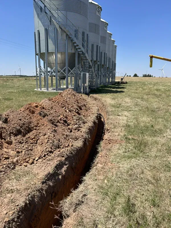 Long trench dug in the ground next to silver grain silos under a blue sky.