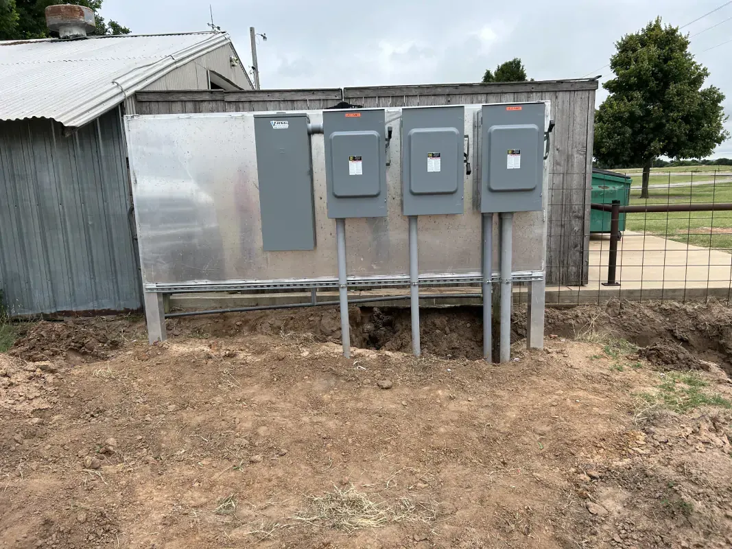 Outdoor electrical panel with three gray boxes, mounted on a metal board. A shed is to the left.