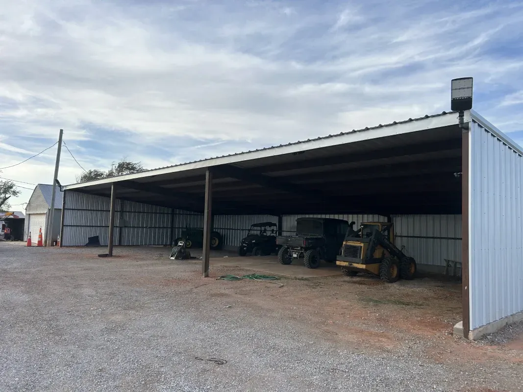 Open-sided metal shed with farm equipment parked underneath, gravel ground, cloudy sky.