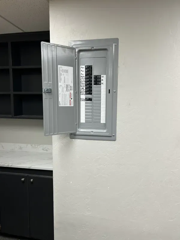 A metal electrical panel with the door open on a white wall next to dark storage cabinets.