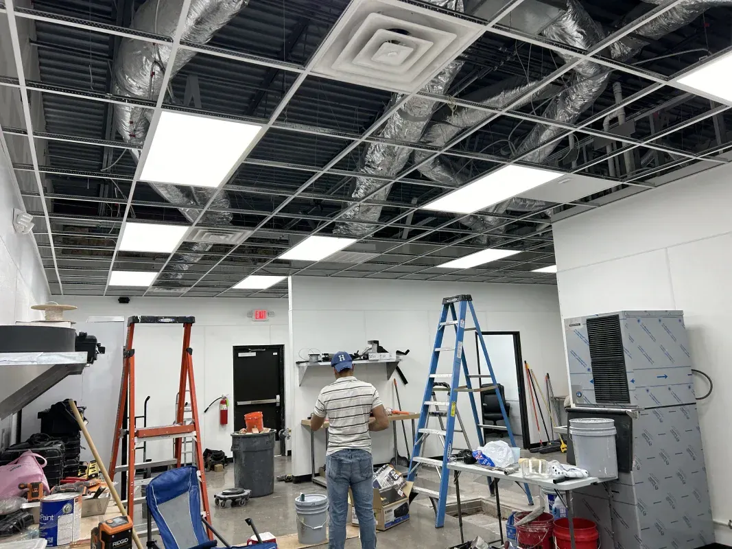 Construction scene: man working on ceiling with ladders, unfinished drop ceiling, ductwork, and lights.