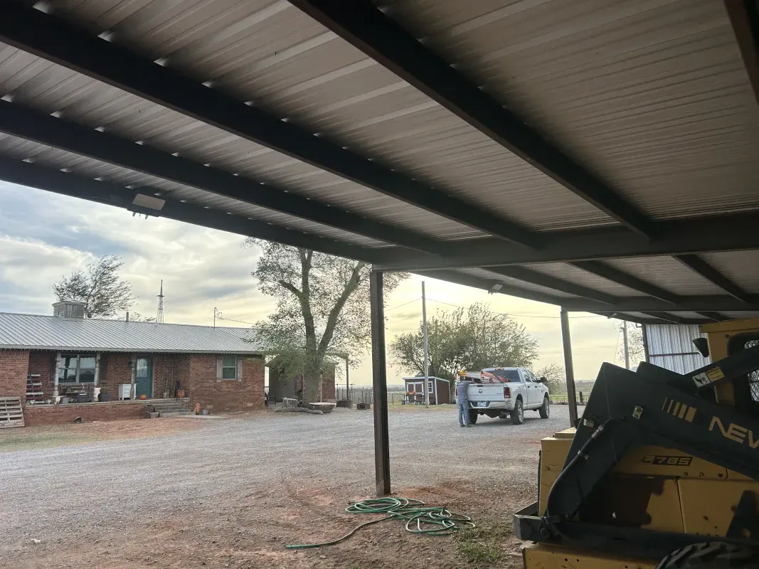 Underneath a metal awning, a gravel yard features a brick house and a white truck parked near trees.