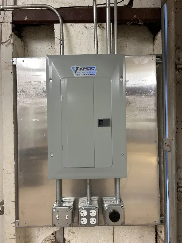 Electrical panel and outlets mounted on a metal plate against a brick wall. Conduit pipes run above and below.