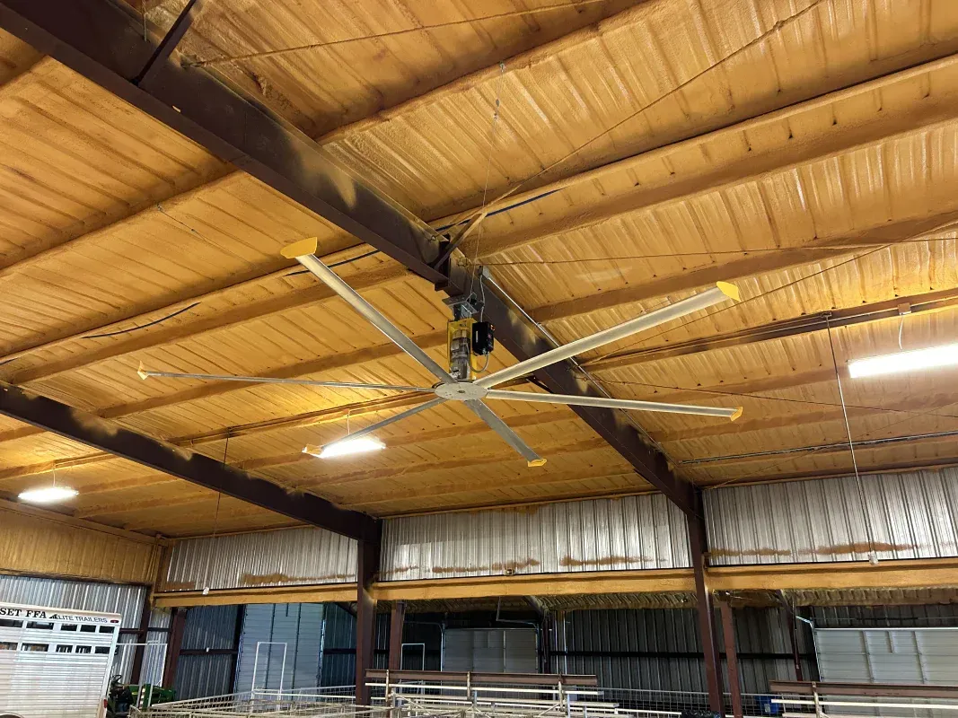 Large industrial fan suspended from the ceiling of a warehouse, with wooden beams and yellow-painted blades.