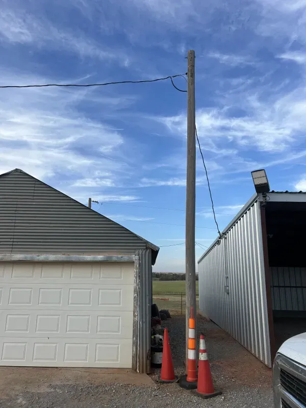 A tall power pole between two metal-sided buildings. Orange traffic cones are at the base. Cloudy sky.