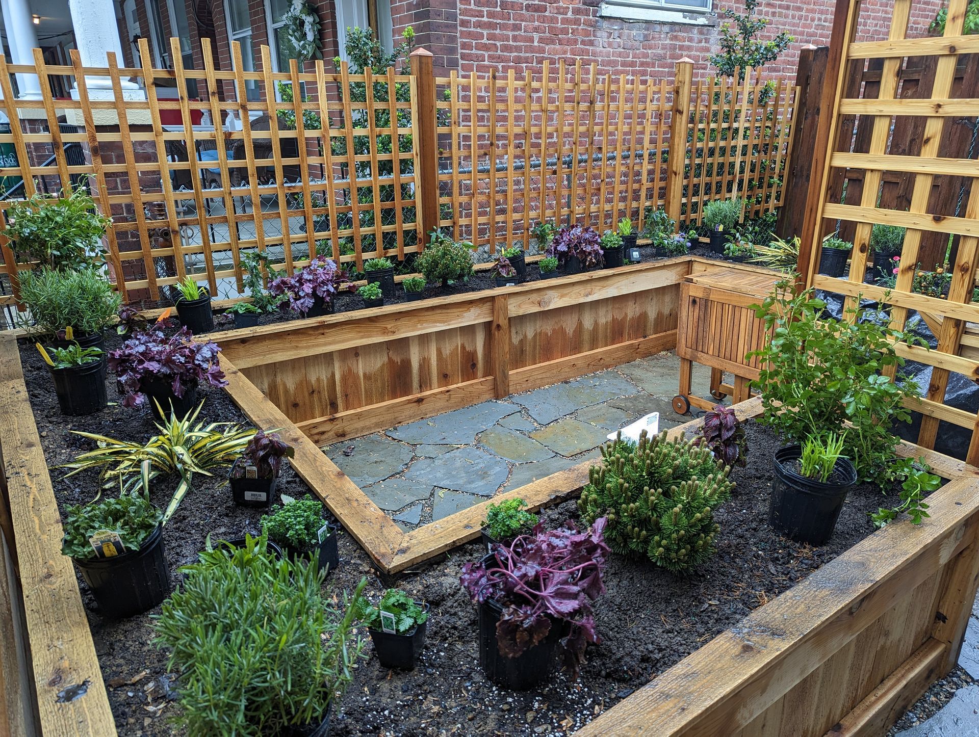 Raised wooden garden bed with lattice, surrounded by potted plants and a brick building.