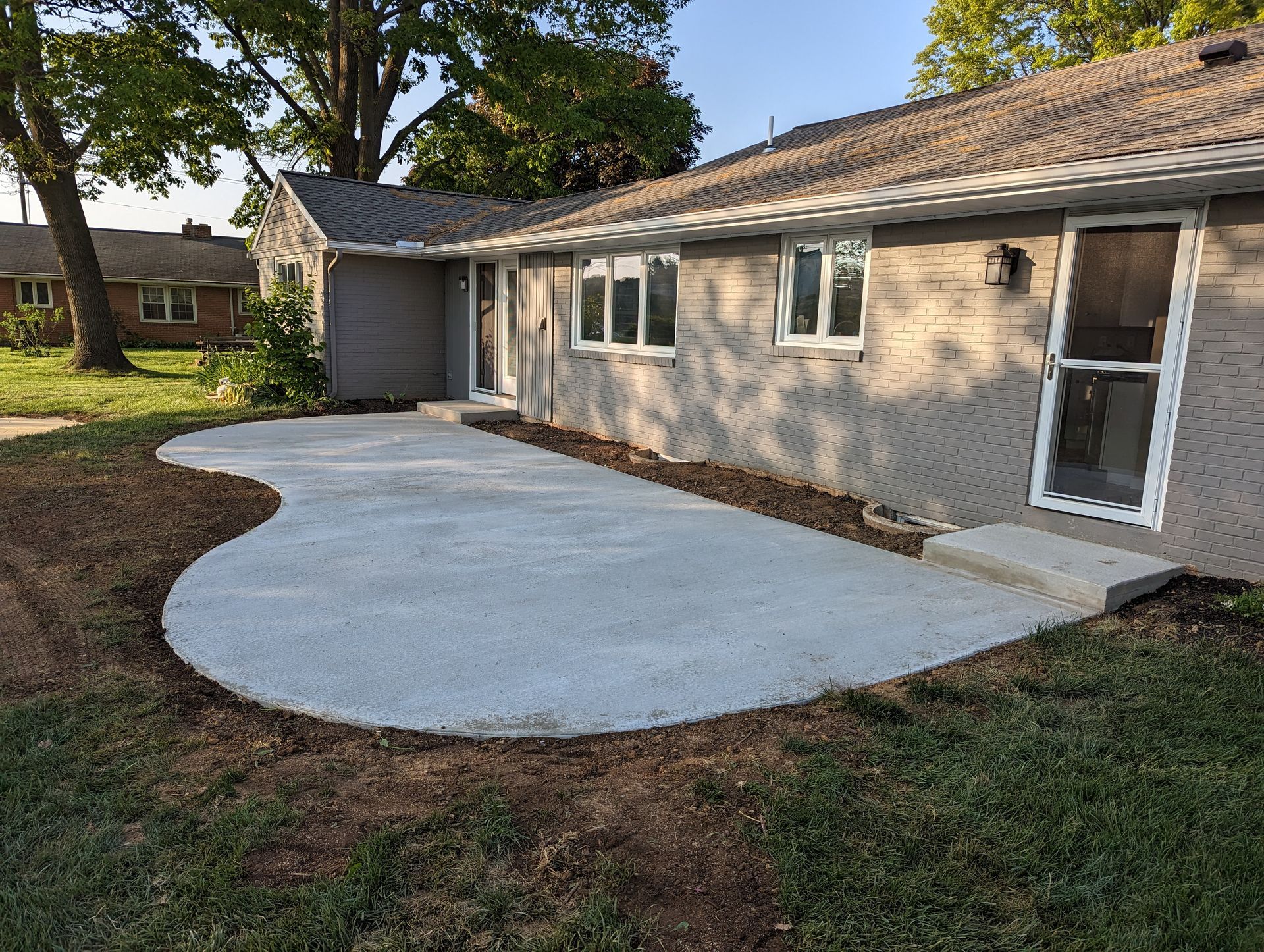 Gray concrete patio next to a light gray house. Newly laid concrete border, grass.