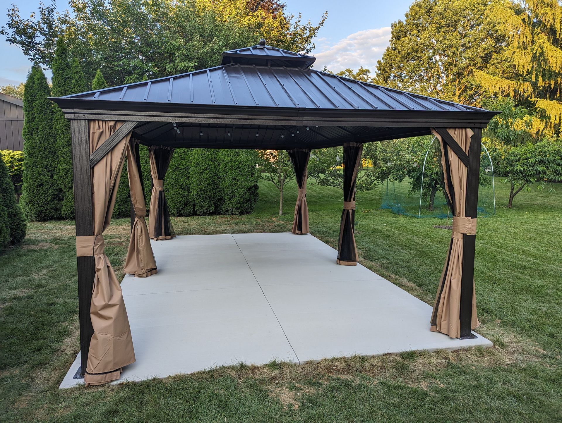Gazebo with brown frame, metal roof, tan curtains, on a concrete patio in a yard with green trees.