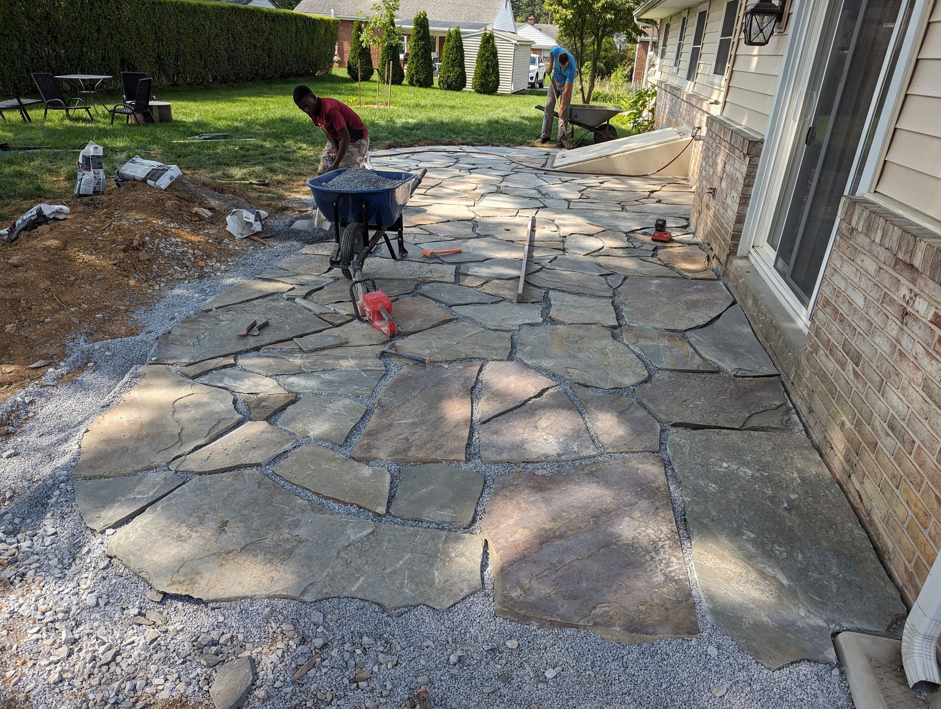 Workers install flagstone patio near a house with gravel base; one uses a wheelbarrow.