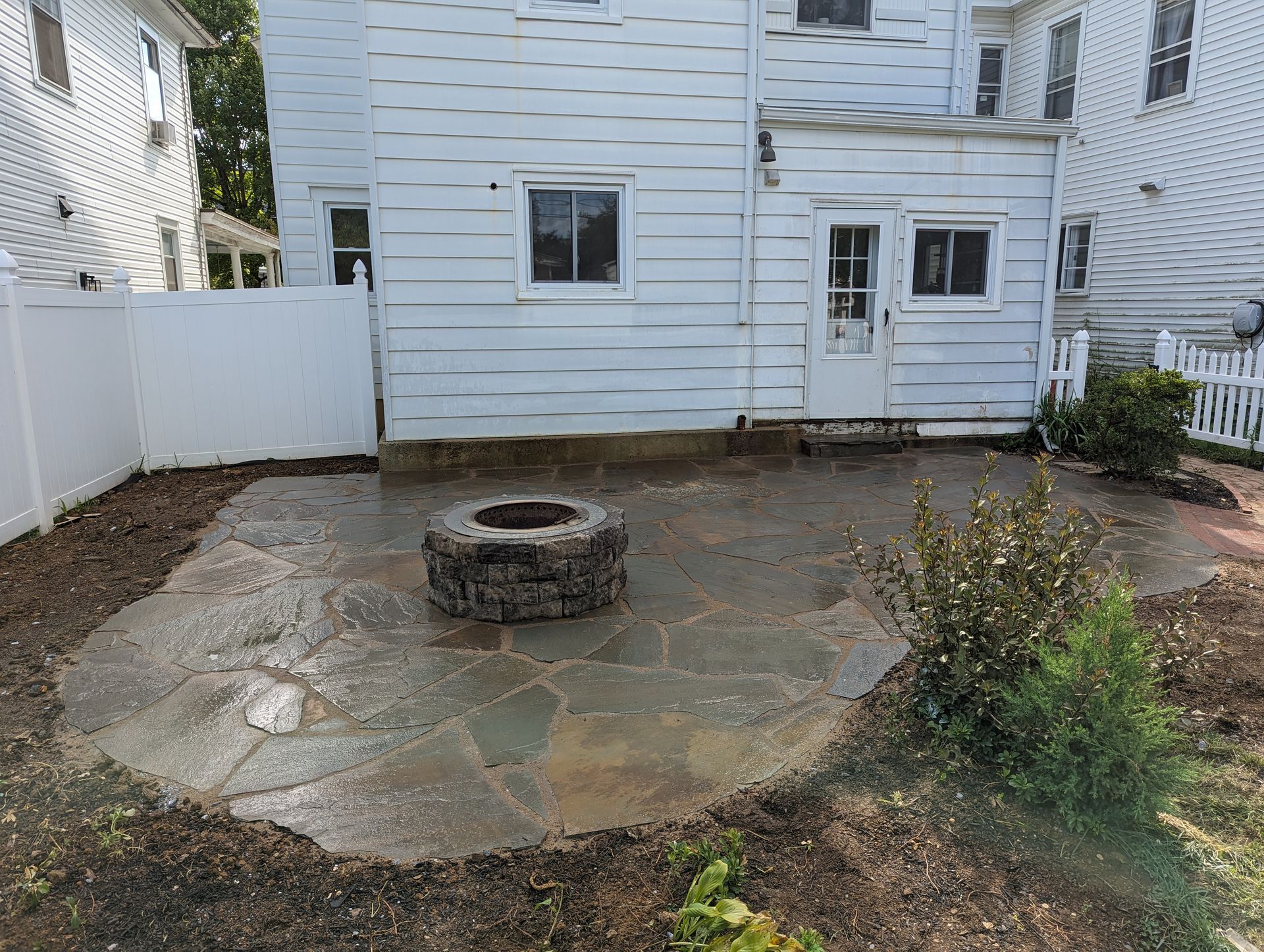 Stone patio with fire pit in front of a white house, surrounded by dirt and landscaping.