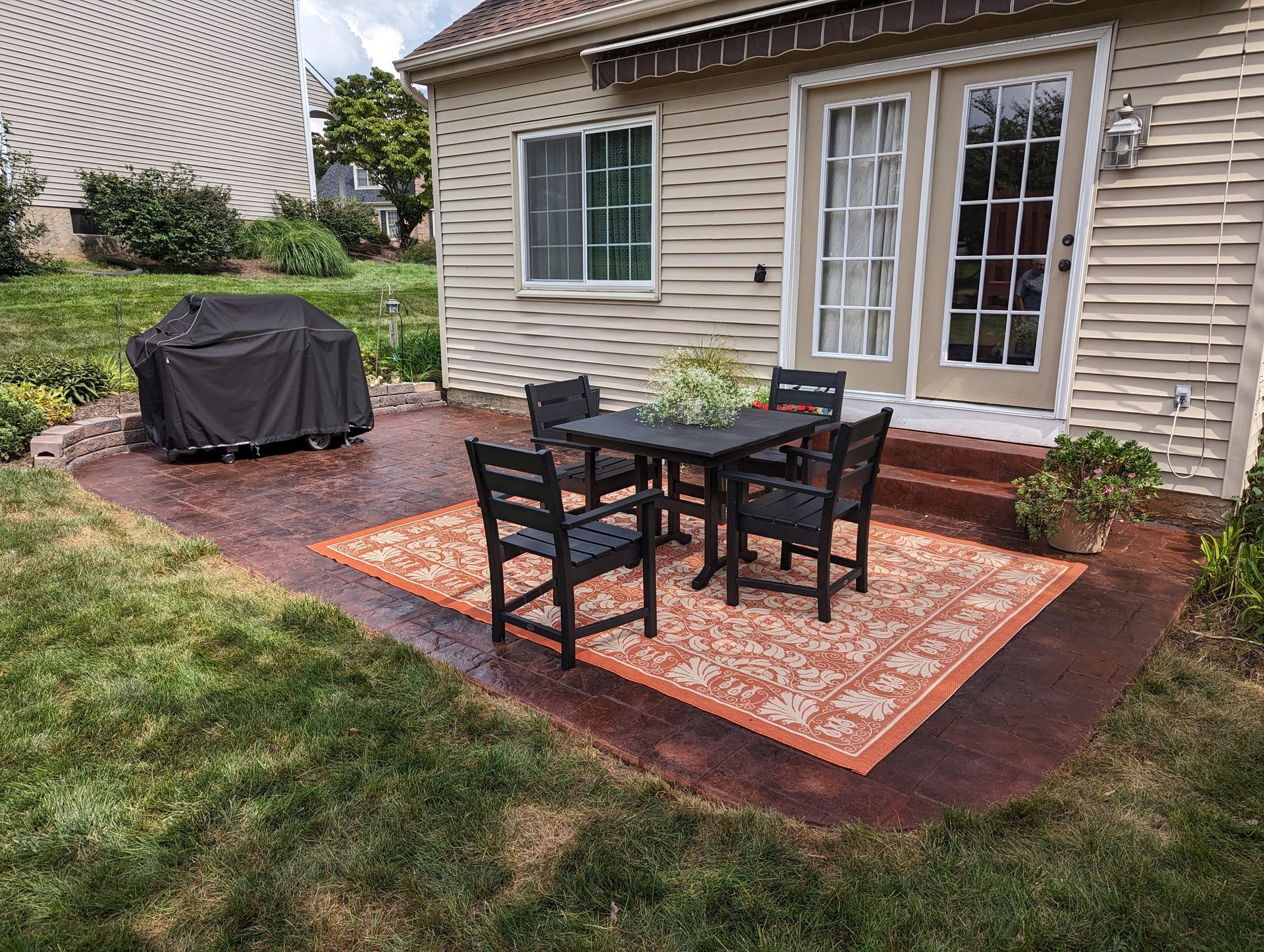 Patio with black table and chairs on orange rug, grill covered, near back door.