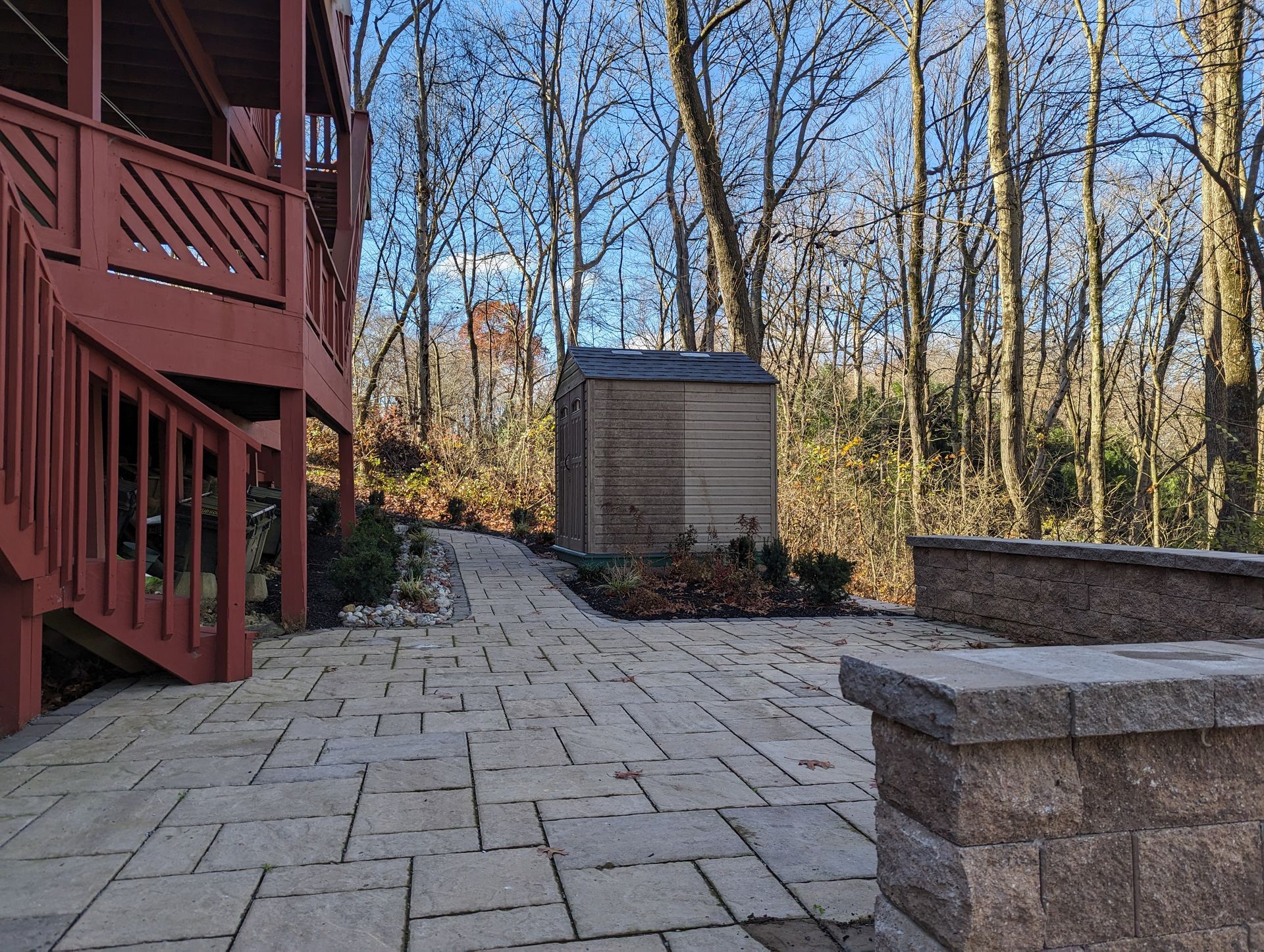Patio with brick pavers, wooden shed, deck, and forest background.