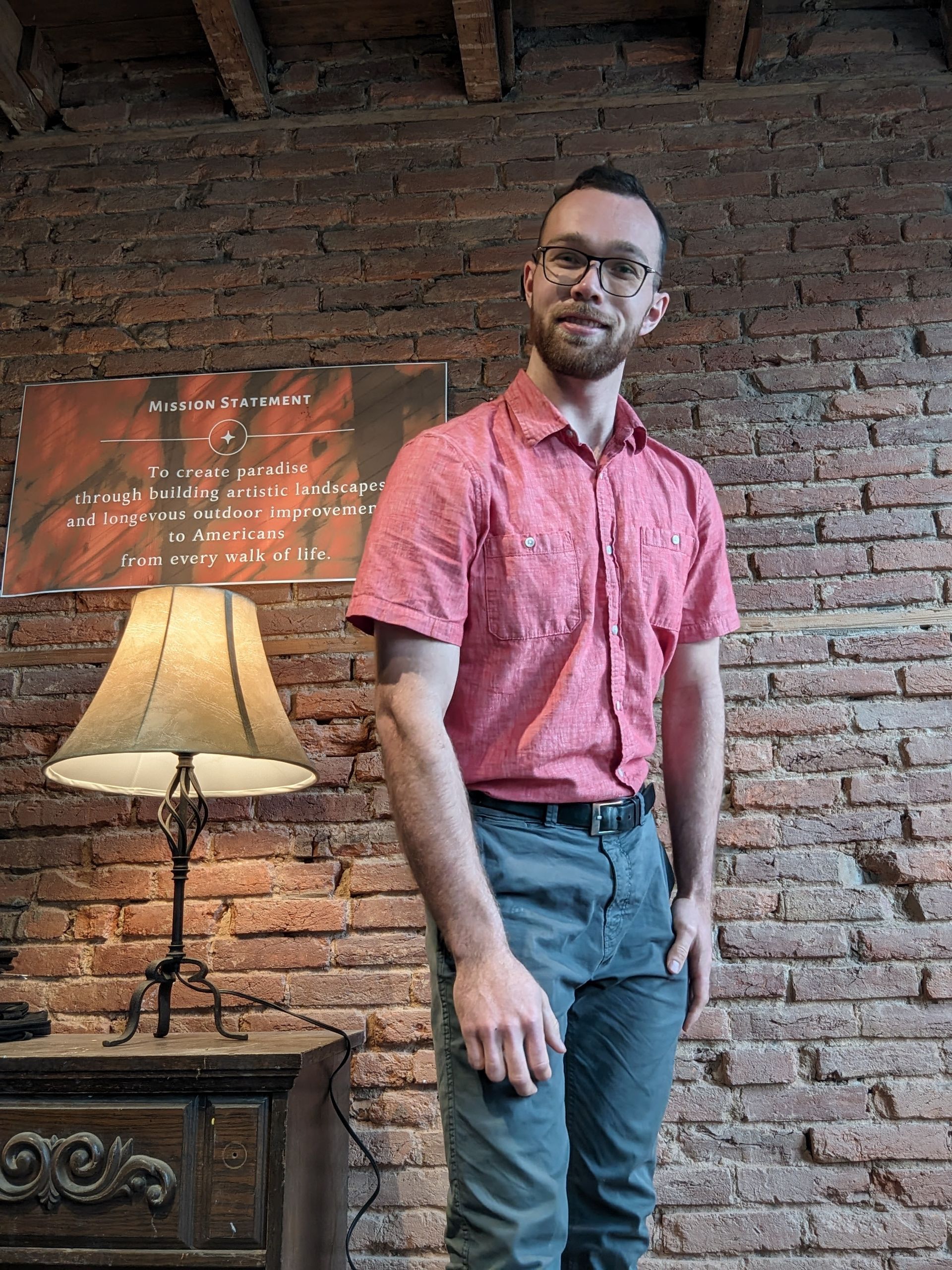 Man in pink shirt and jeans stands by brick wall, next to a lamp and ornate table.