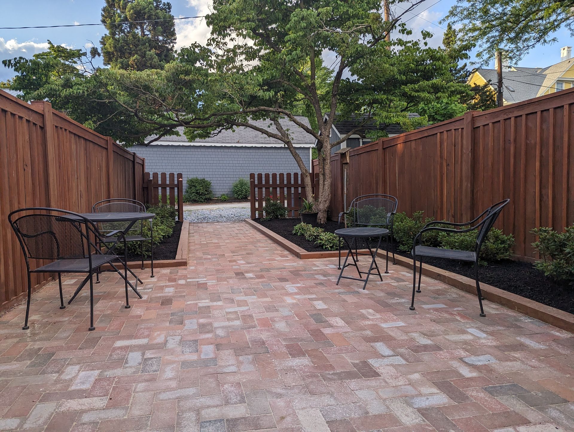 Brick patio with black wrought-iron table and chairs, enclosed by brown wooden fence and trees.