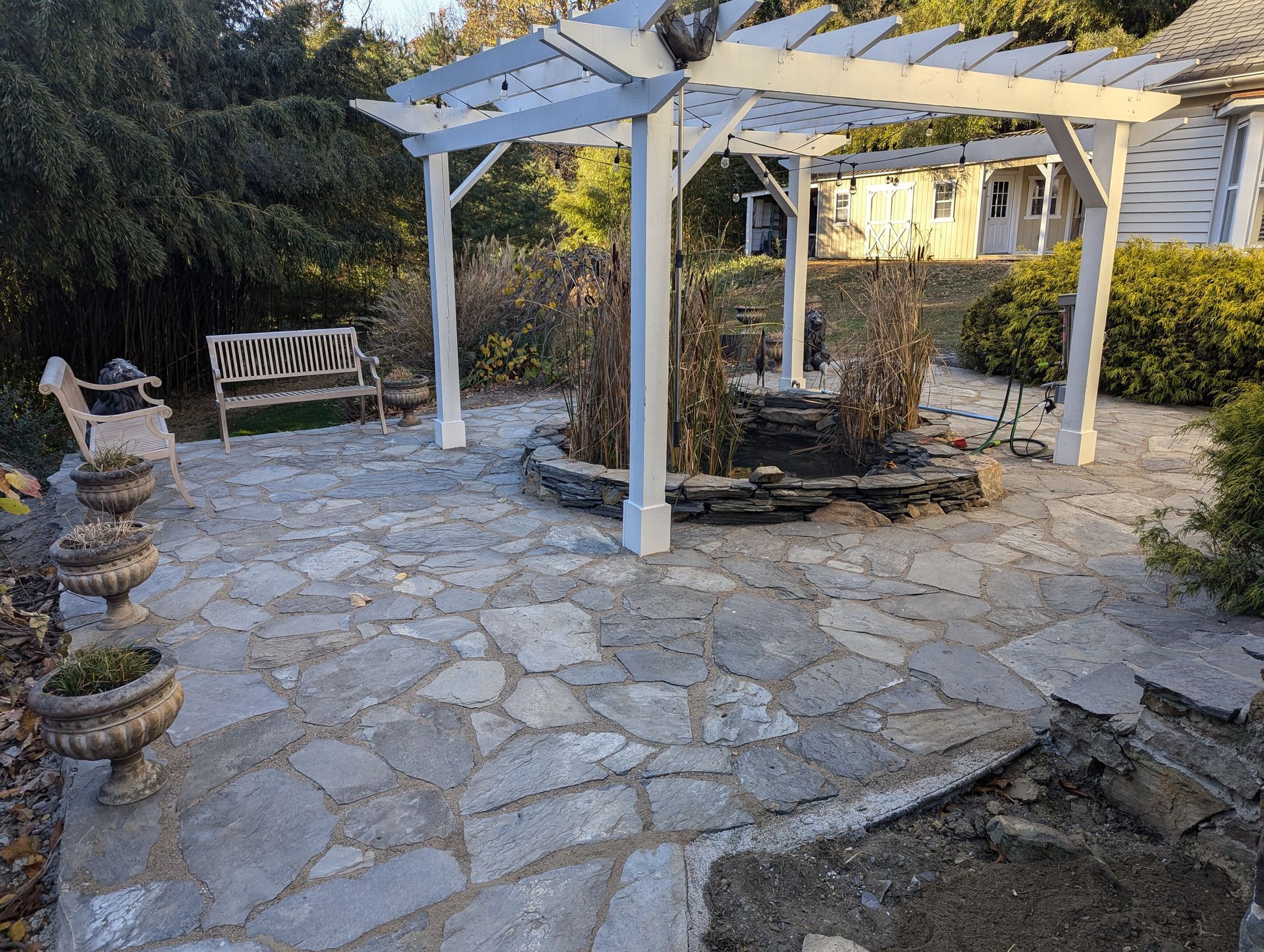 Stone patio with white pergola, bench, and planters in a garden setting.