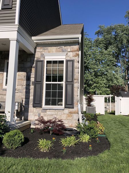Stone facade with a window and black shutters, bordered by a garden and a white fence.