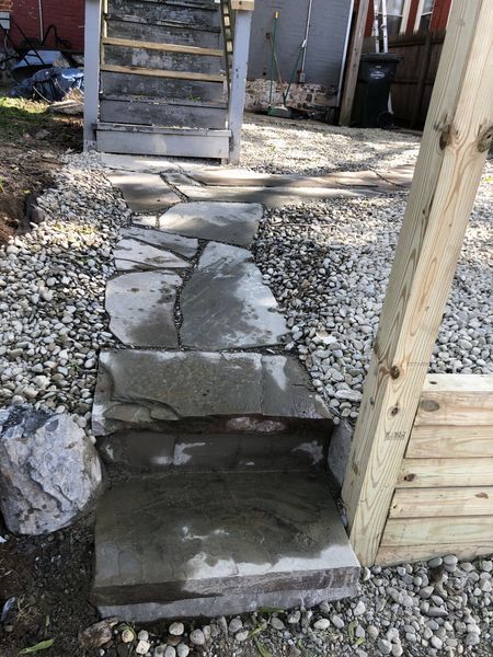 Stone pathway leading to wooden stairs, next to a wooden structure, set on gravel.