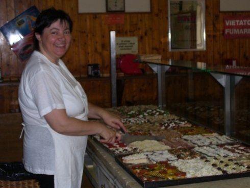 una donna dietro a un bancone mentre farcisce delle pizze in teglia