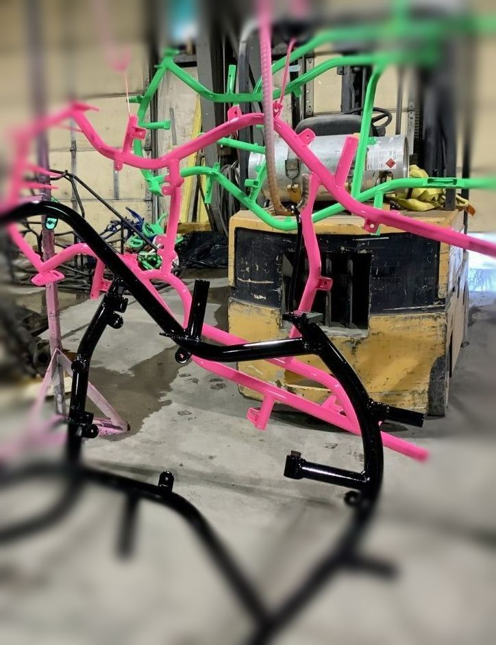 Pink and green metal frames drying on a forklift in a workshop; a black frame is in the foreground.