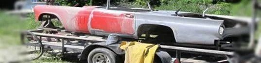 A car frame, half red and half gray, sits on a trailer in a grassy area.