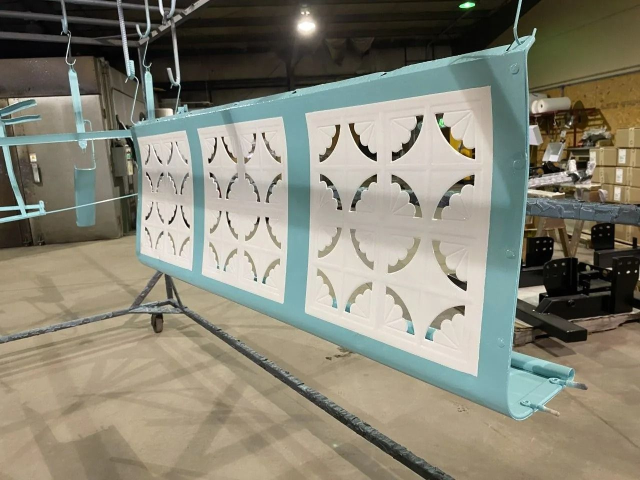 A large, teal metal panel with three white, decorative cut-out sections, hanging in a workshop.