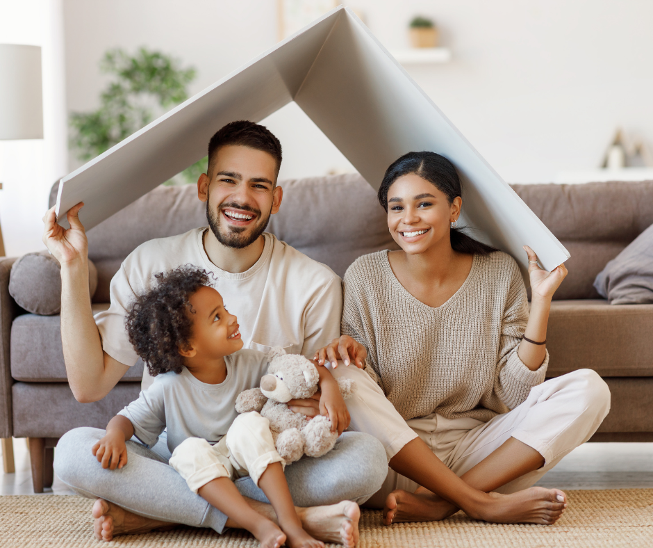 A family is sitting on the floor under a cardboard house.