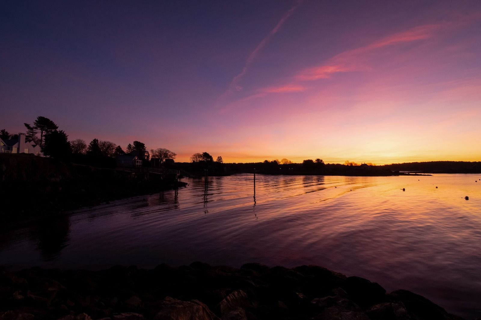 Sunset over calm water with purple, pink, and orange hues. Silhouette of trees and shore.