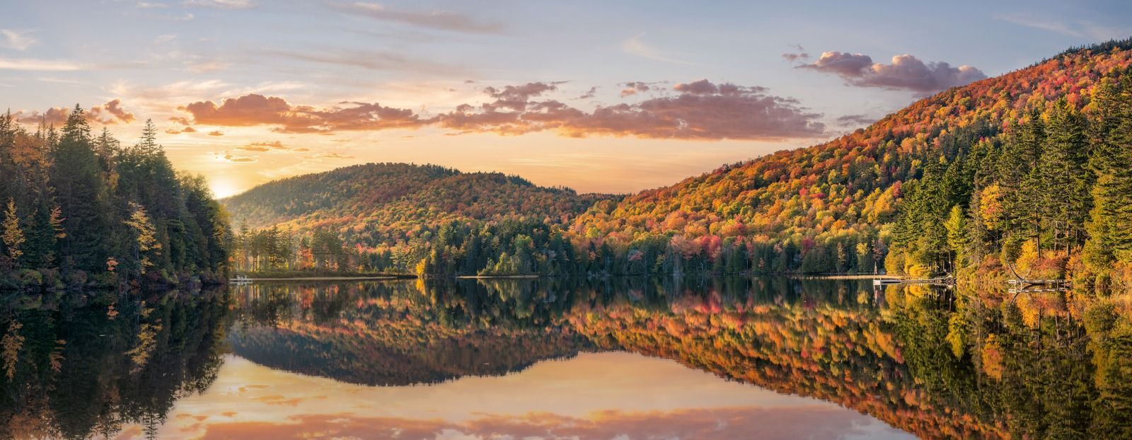 Autumn landscape with mountains, colorful trees reflected in a calm lake, and a bright sky.