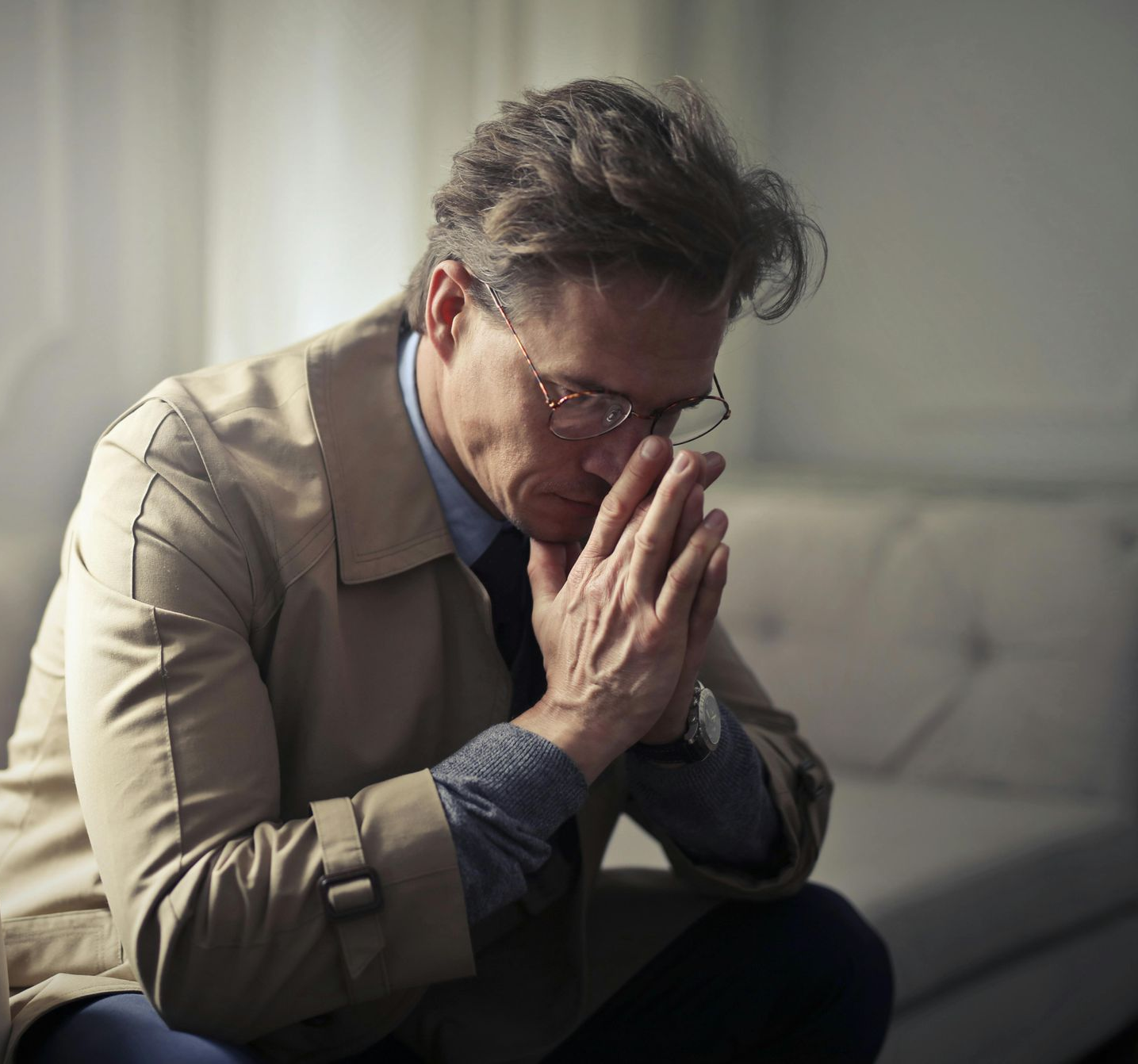 Man in glasses, trench coat, hands clasped, appears pensive. Sitting indoors, soft lighting.