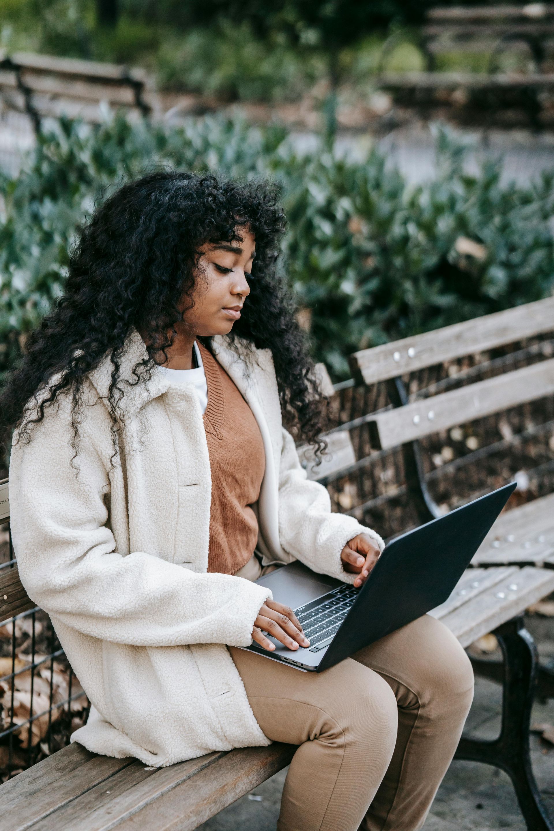 Woman using a laptop on a park bench, wearing a cream coat, brown sweater, and pants.