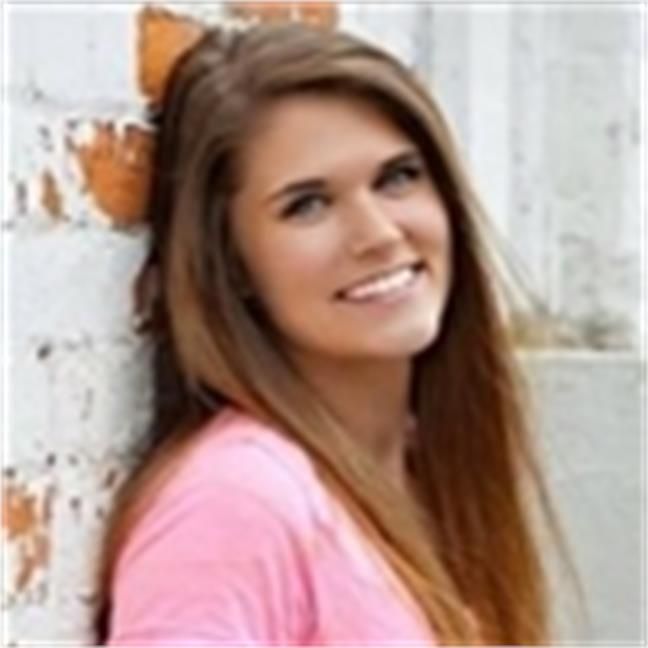 A woman in a pink shirt is leaning against a brick wall and smiling