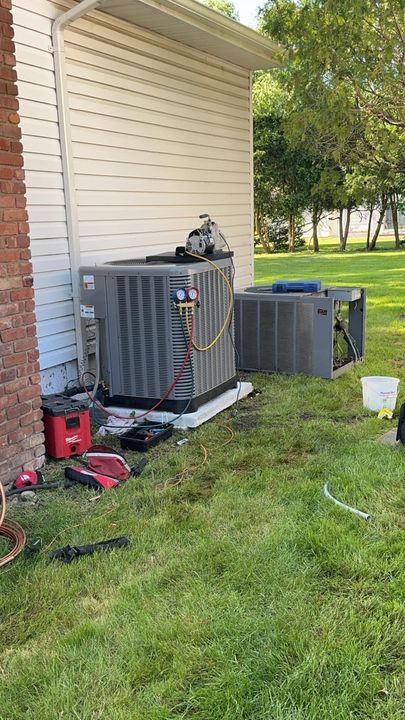 Two outdoor HVAC units beside a house wall, with tools and a vacuum on the grass.