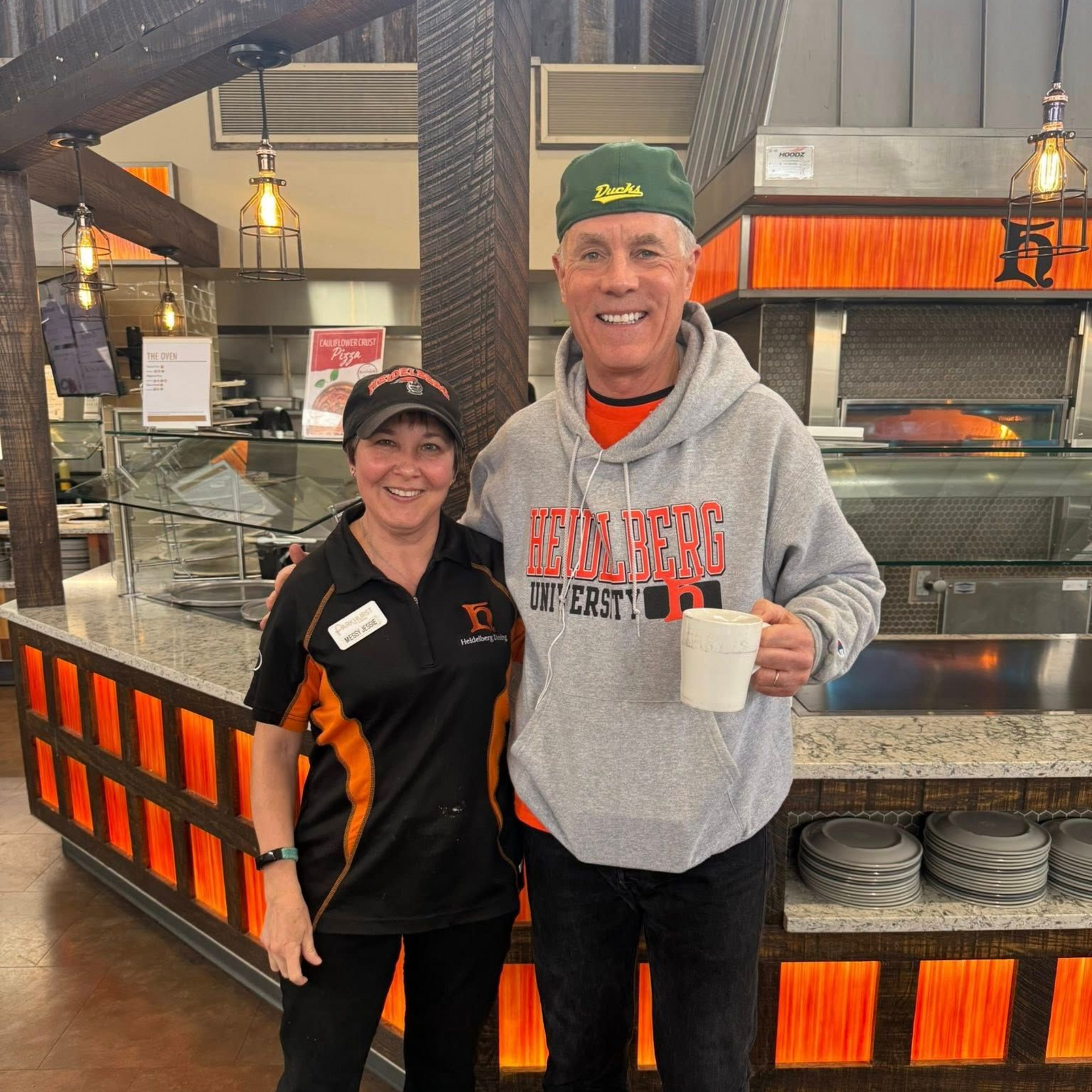 Woman in uniform and man in a hoodie pose together in a restaurant. The man holds a mug.
