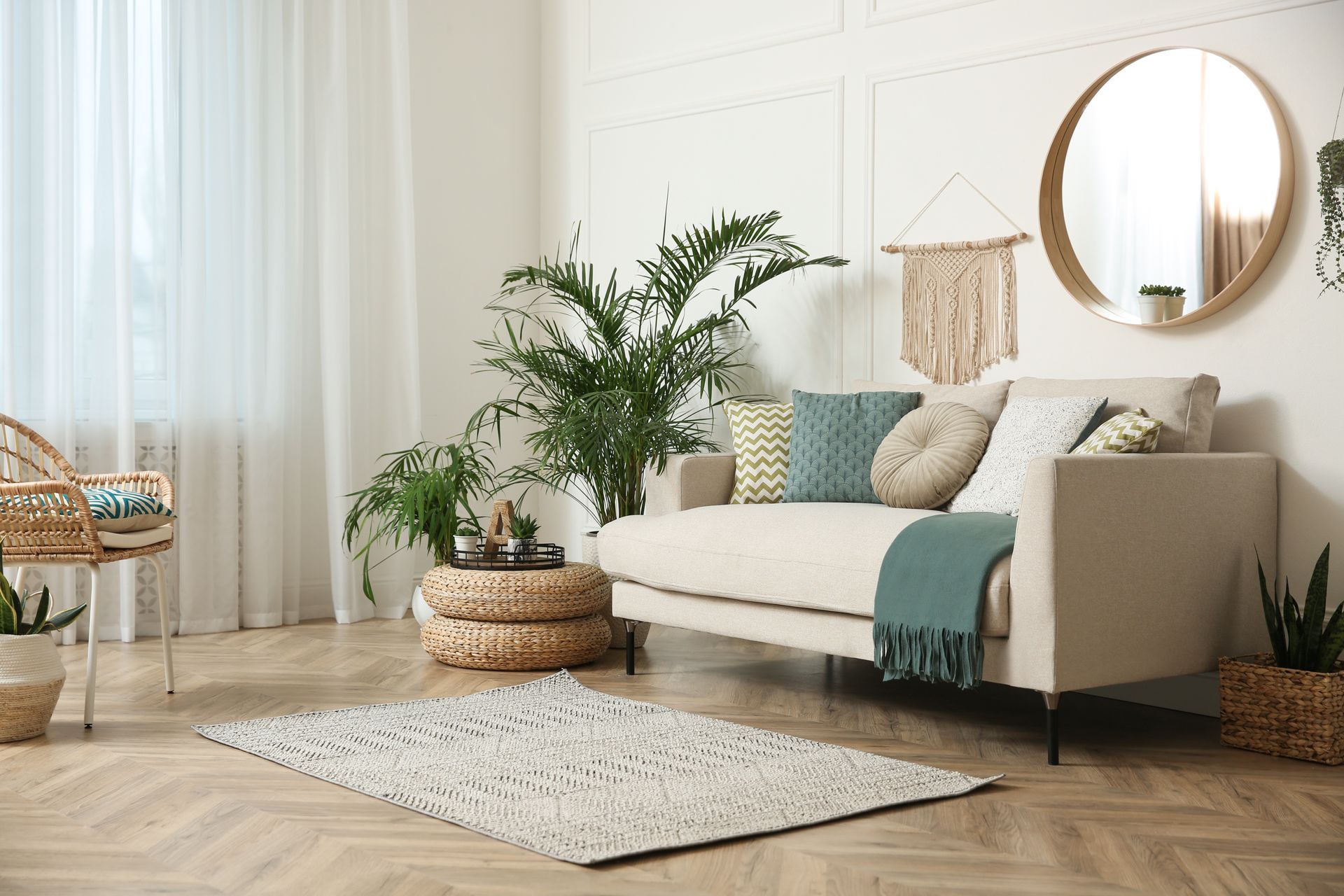 Living room with a beige sofa, round mirror, plants, woven rug, and light-colored walls and floors.