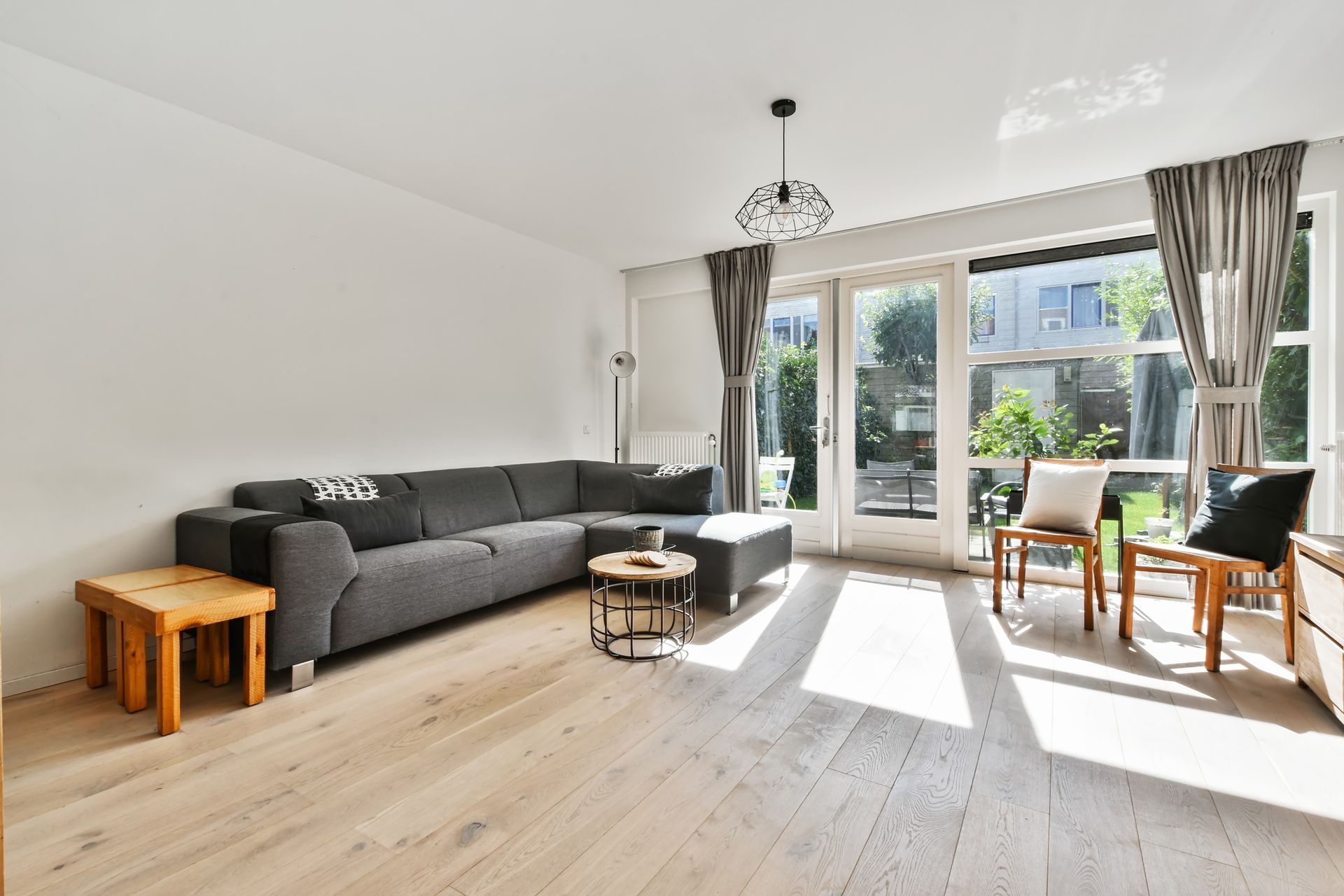 A modern living room with a gray sectional sofa, light wood floors, and large windows with gray curtains.