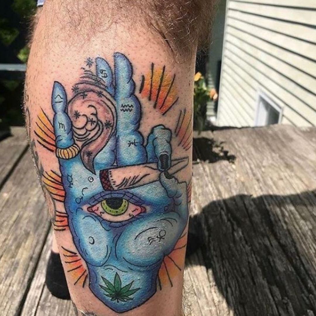 Blue hand tattoo holding a lit joint with an eye on the palm and a cannabis leaf.