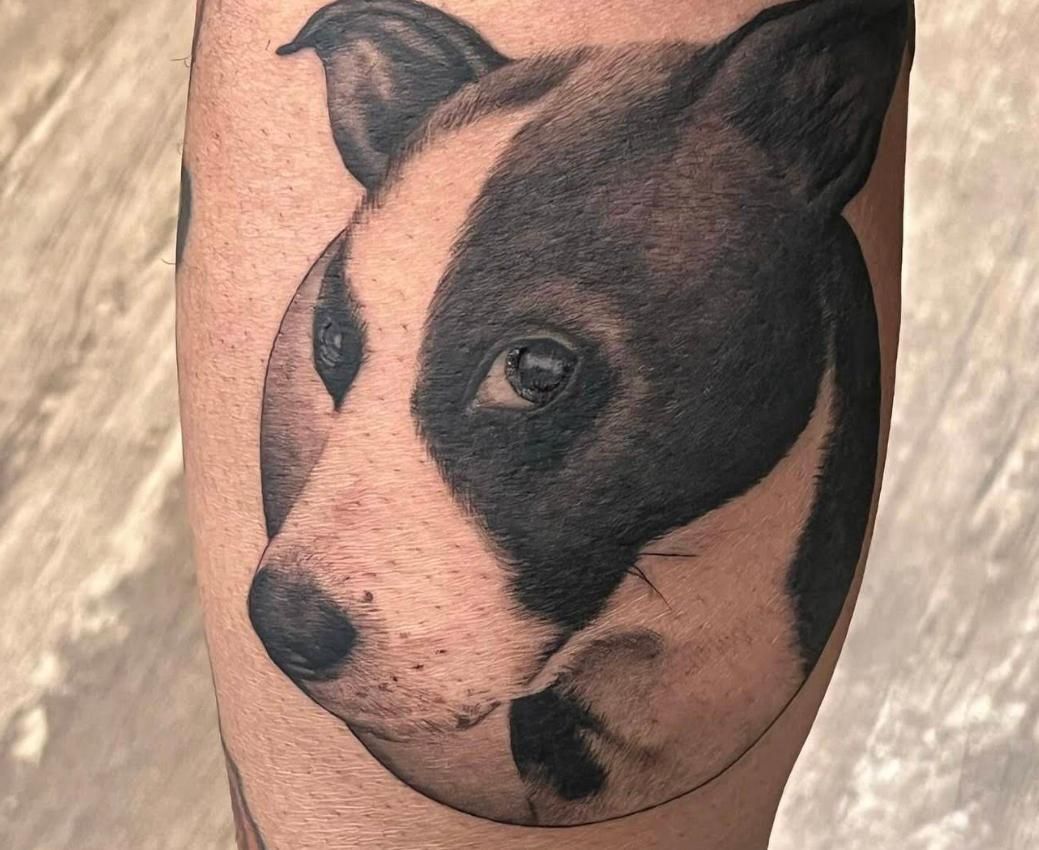 Black and white tattoo of a dog's head on skin; one eye visible.