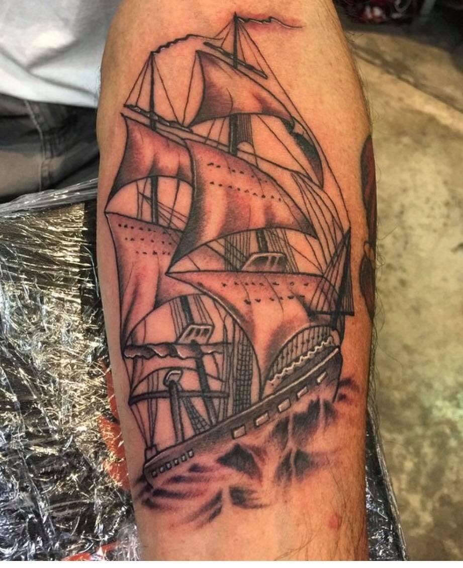 Tattoo of a sailing ship with billowing sails on a forearm; waves at the base, grayscale.