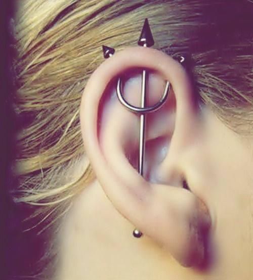 Ear with a trident industrial piercing, silver jewelry, and blonde hair.