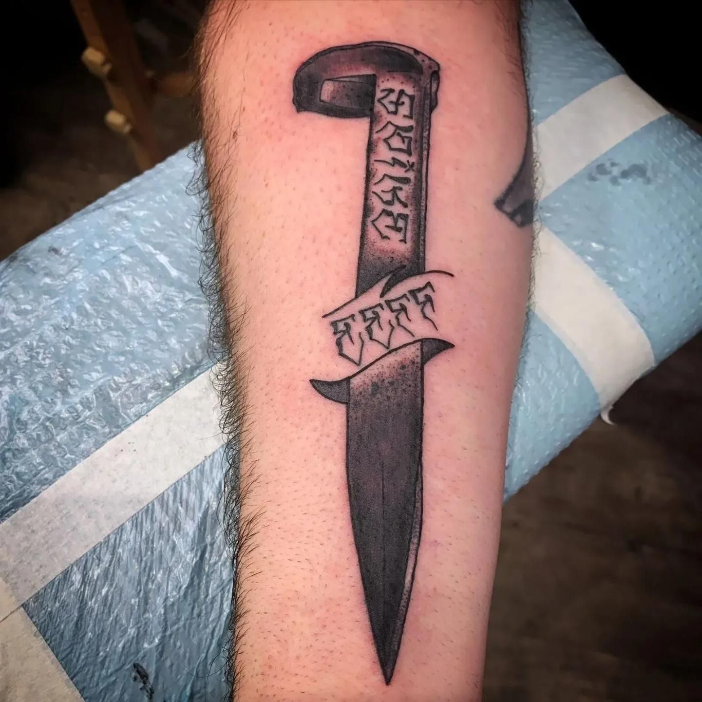 Tattoo of a dagger with text on an arm, black and grey ink on a blue surface.