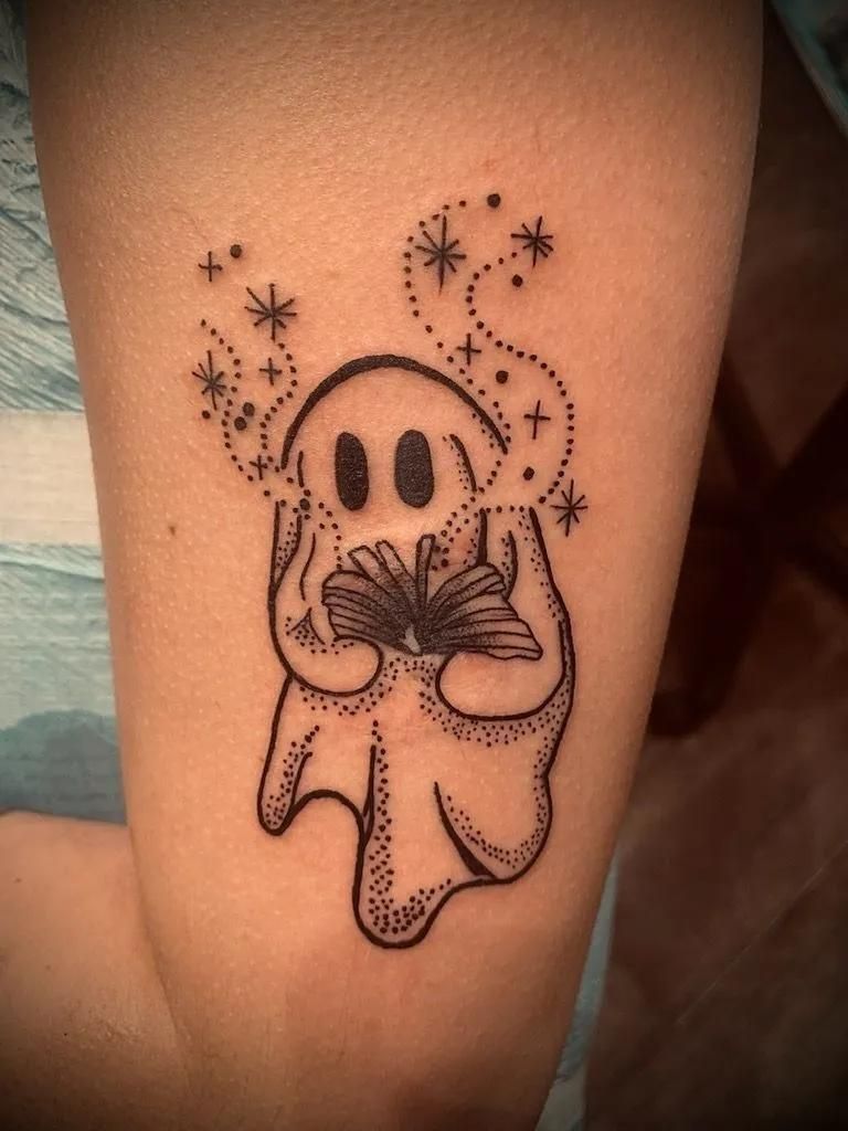 A tattoo of a ghost holding a book, with stars and dots emanating from the book.