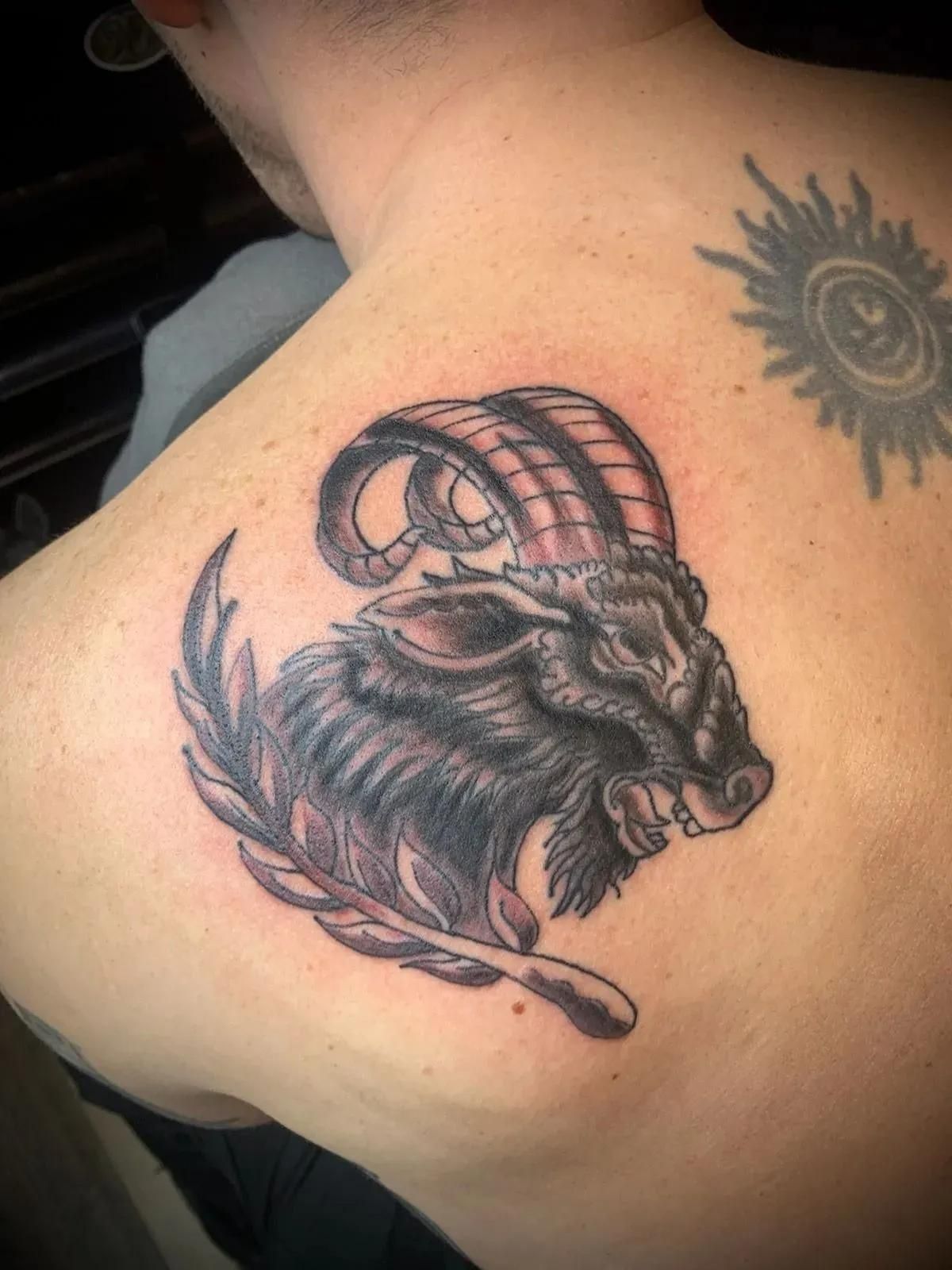 Tattoo of a ram's head with large curved horns and laurel branch on a person's back.