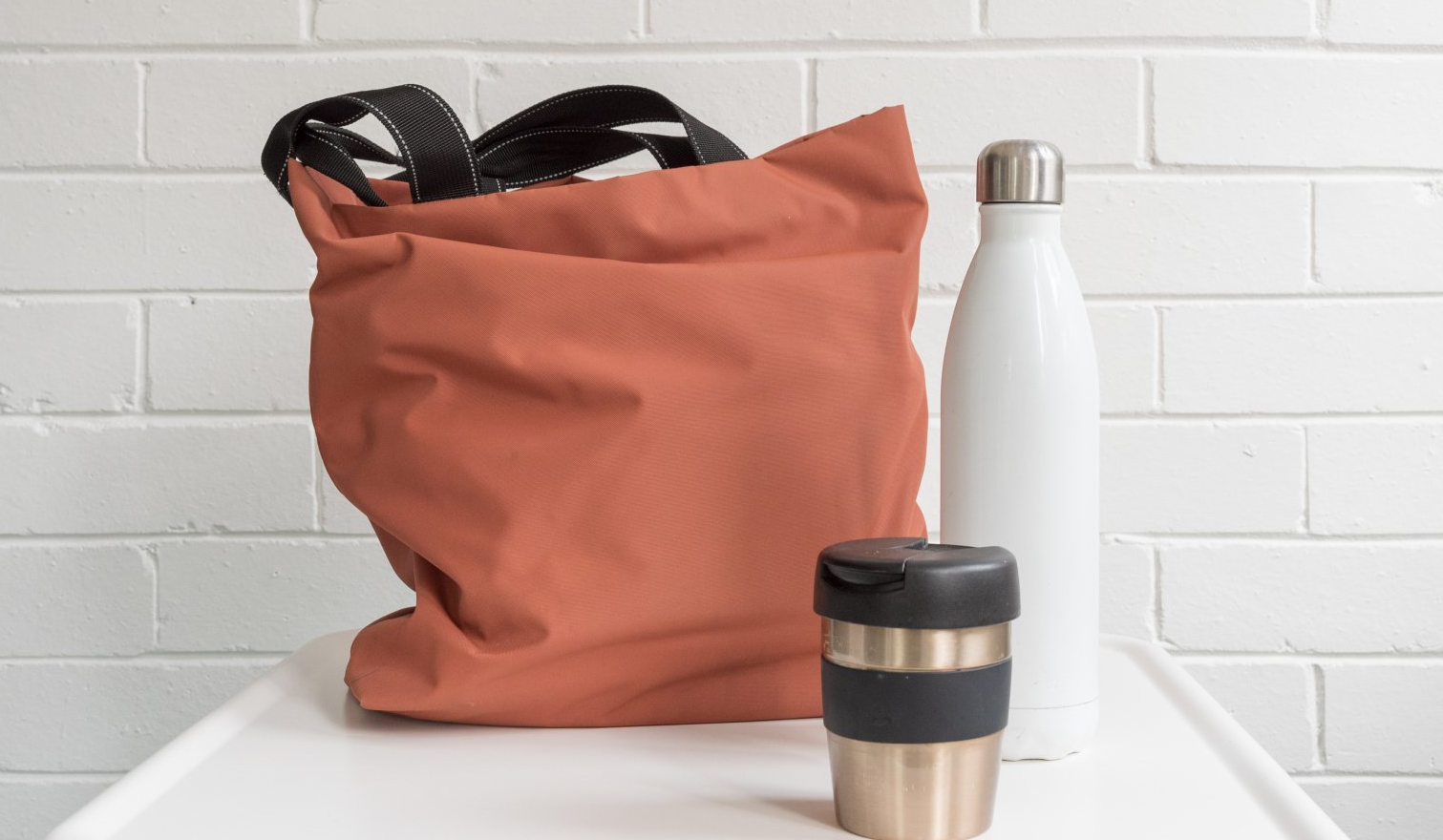 A tote bag , a bottle , and a cup are on a table.